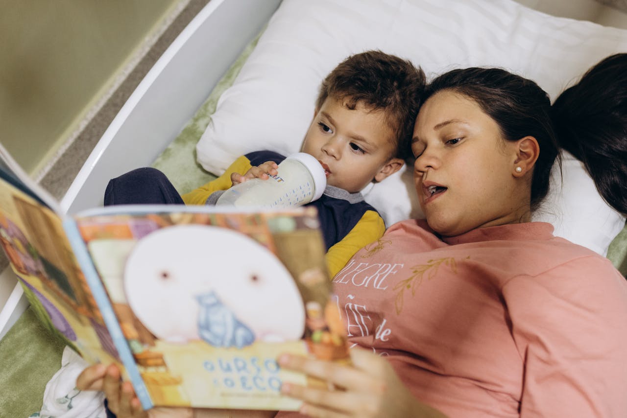 gallery-1 A mother reads a bedtime story to her child while he drinks milk, creating a cozy and bonding moment.