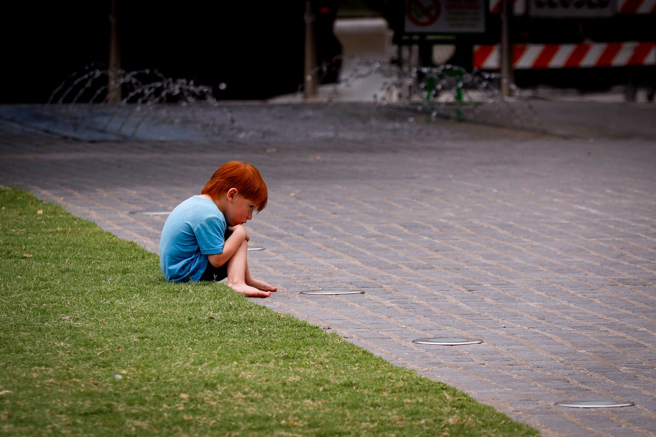 A young red-haired child sits thoughtfully on the pavement near a grassy area in an urban setting.