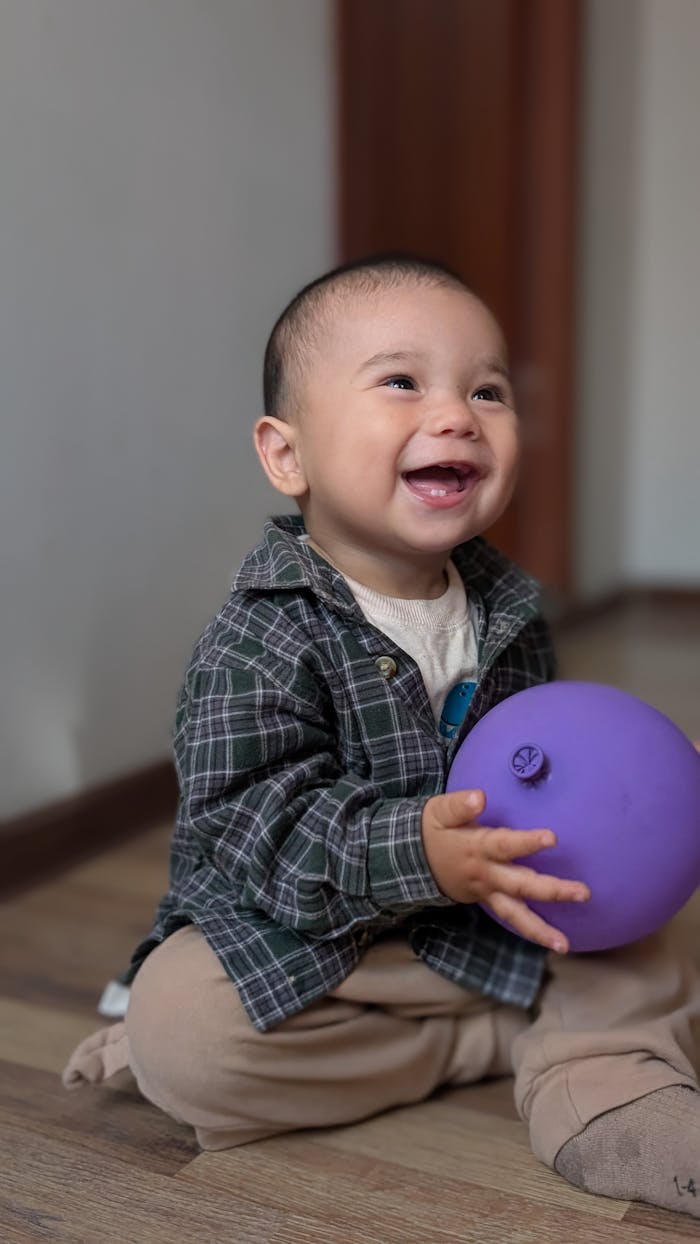 services-01 Happy baby in plaid shirt playing with a purple balloon indoors on a wooden floor.