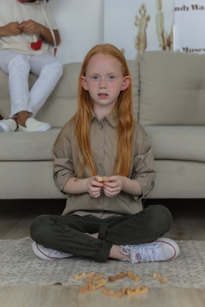 Full body of cute red haired child in stylish clothes sitting on floor and playing game while looking at camera