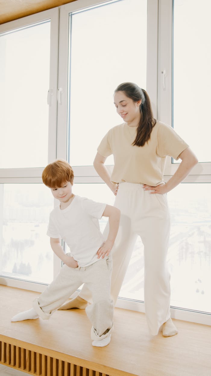 gallery-4 A woman and child practicing yoga poses indoors by a sunny window, promoting family fitness.