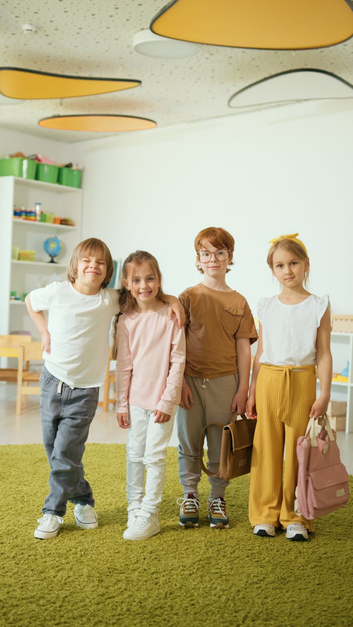 gallery-6 Four cheerful children standing together, smiling in a classroom environment with bright decor.