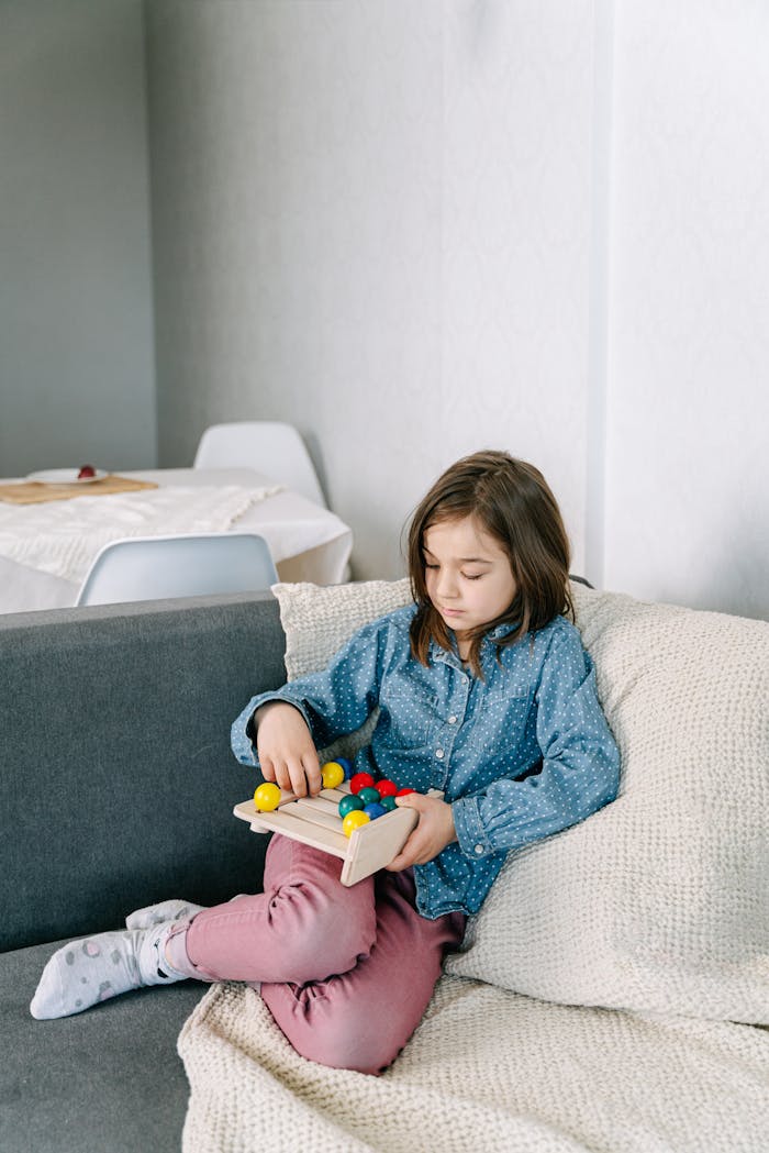contact-img Young girl engaged in mindful play with wooden toy on sofa, indoors.